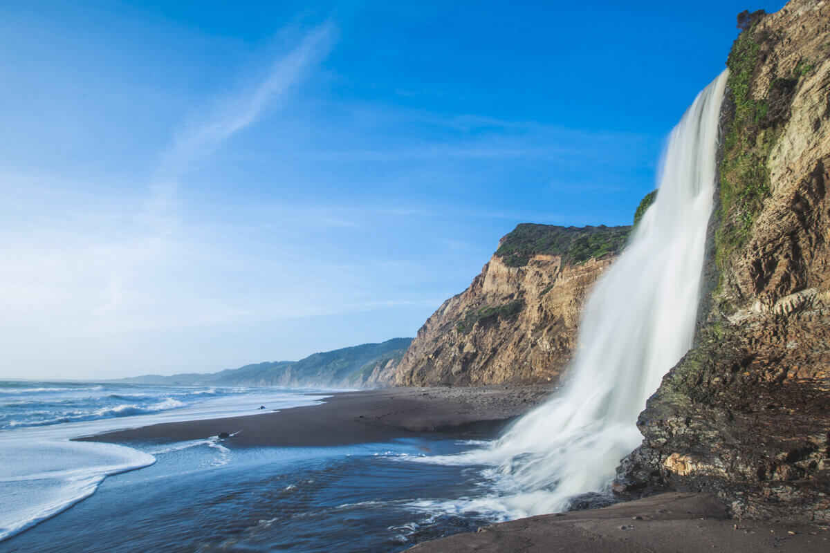 Alamere Falls cascading down from the cliffs onto the beach right besides the ocean on a sunny day.