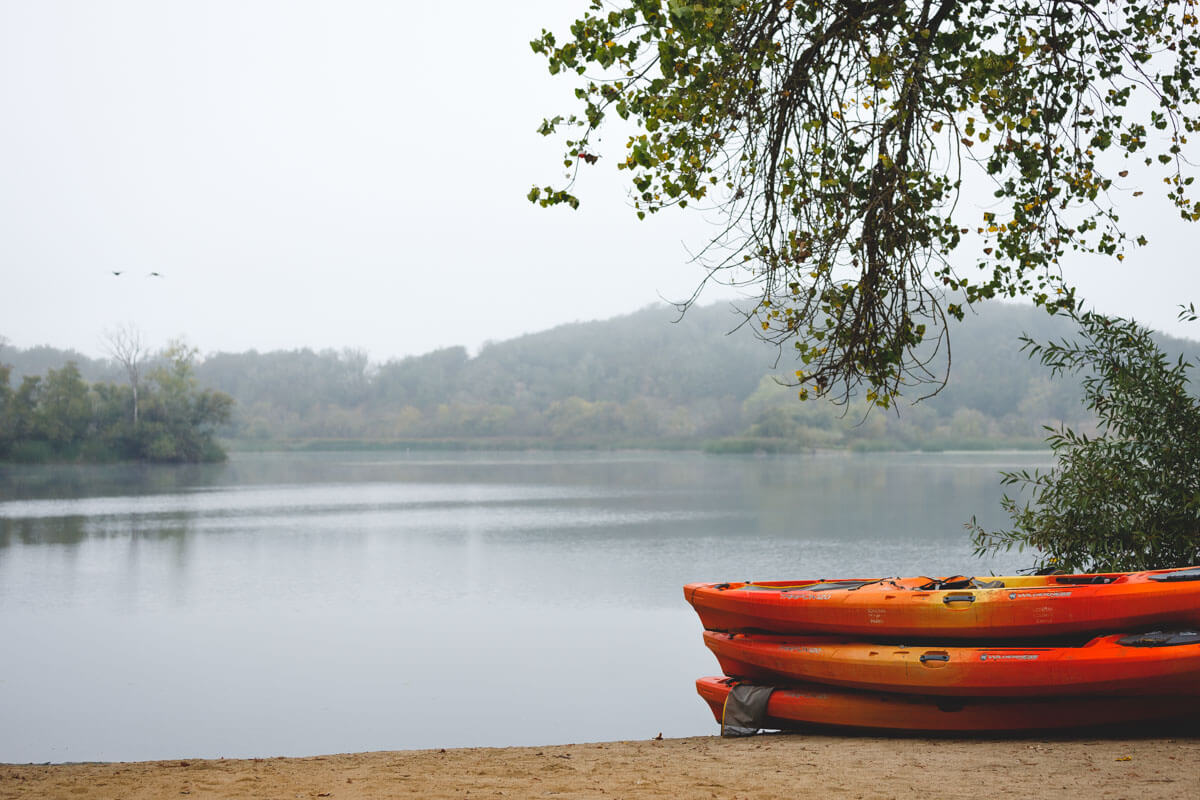 Red kayaks piled up on a beach beside Spring Lake with a view across to forests on an overcast day.