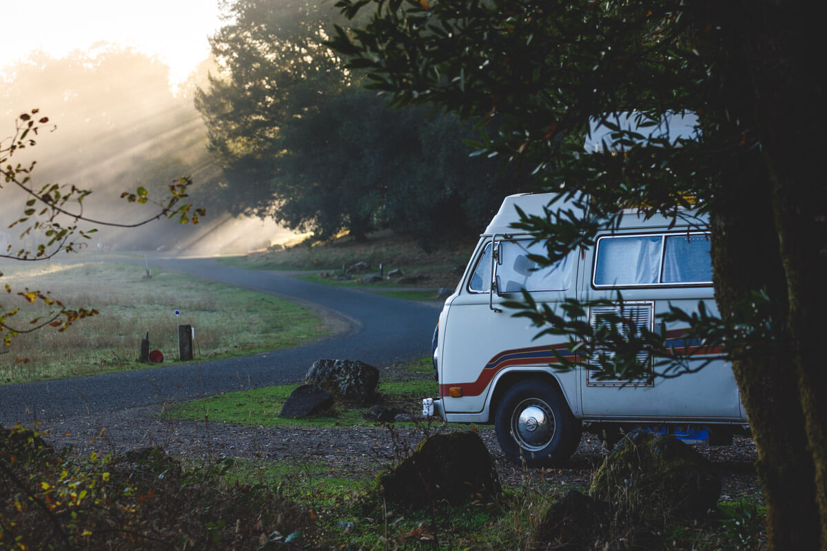 A white campervan at Sugarloaf Ridge State Park Camping in between trees while golden hour light rays shine down.