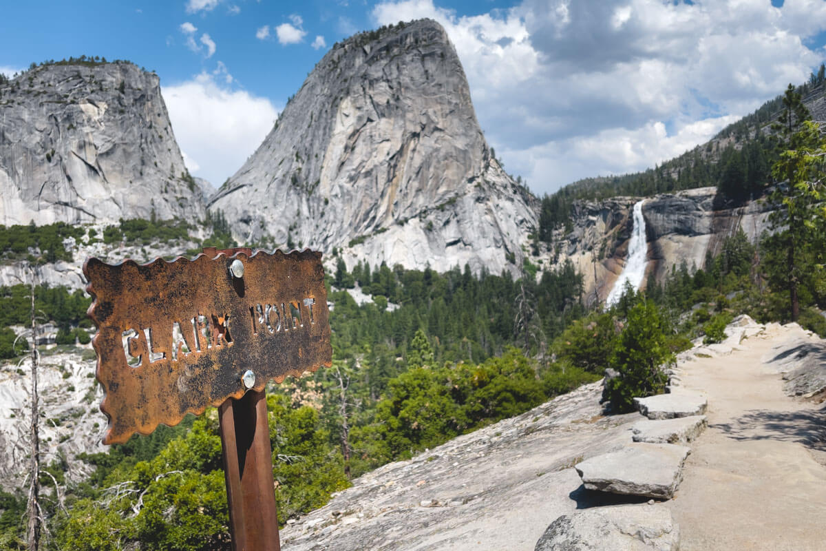A very rusty sign for Clark Point trail with the path leading to Nevada Falls in the distances sandwiched by the mountains of Yosemite.
