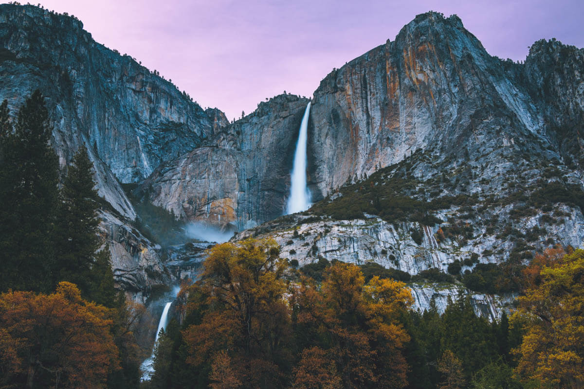 Fall-colored trees surrounding the huge Yosemite Falls in Northern California with a stunning sunset sky.