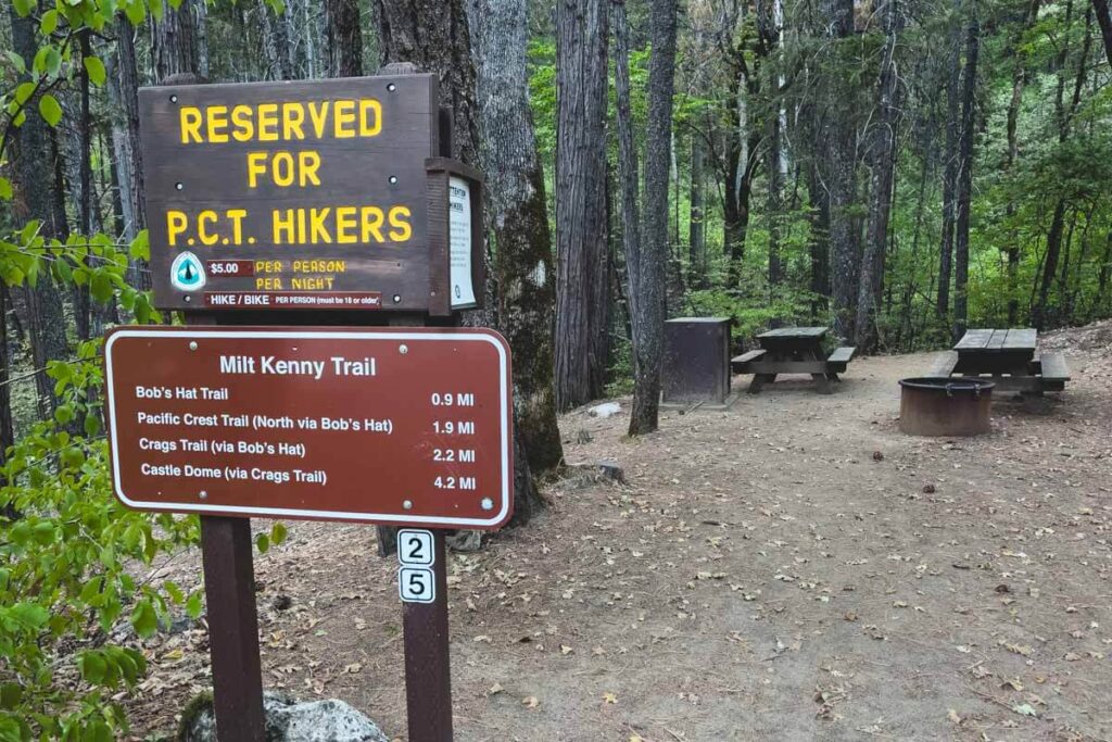 The Milt Kenny trailhead sign, with other trail information, at the entrance to a campsite with a BBQ pit and multiple picnic benches.