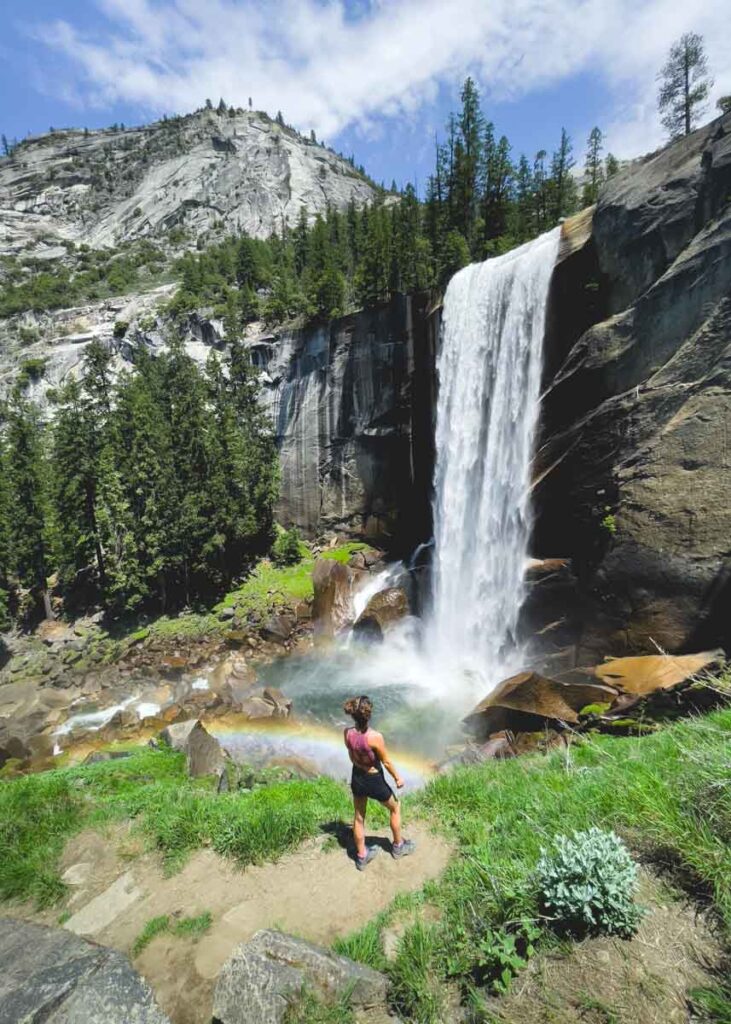 A lone hiker stands in front of Vernal Falls in Yosemite National Park while the waterfall produces a rainbow.