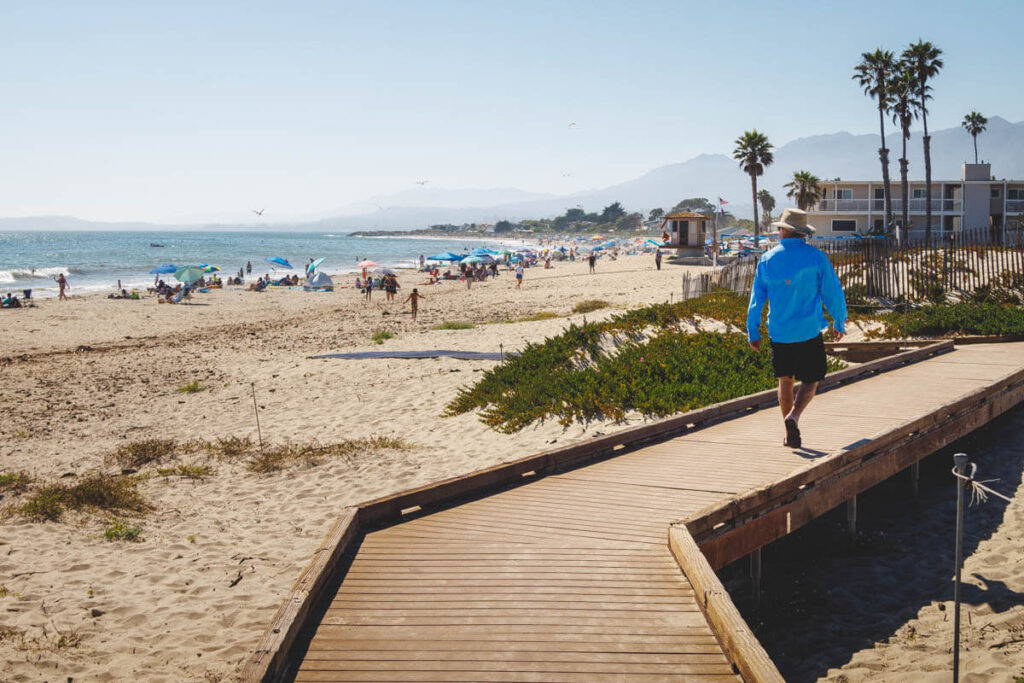 A man walking along the wooden boardwalk next to Carpinteria State Beach on a sunny day.