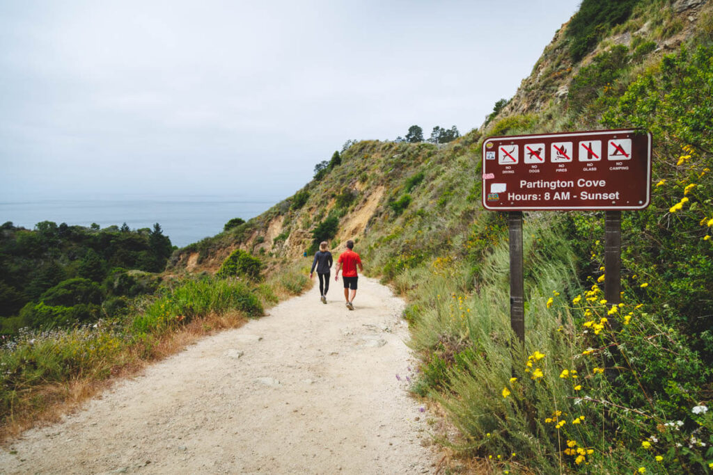 Two tourists walking along Partington Cove trail besides the trailhead sign.