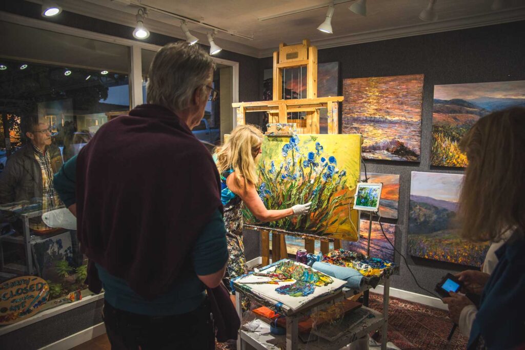 A female artists paints and oil painting in Carmel-by-the-Sea art gallery while people watch.