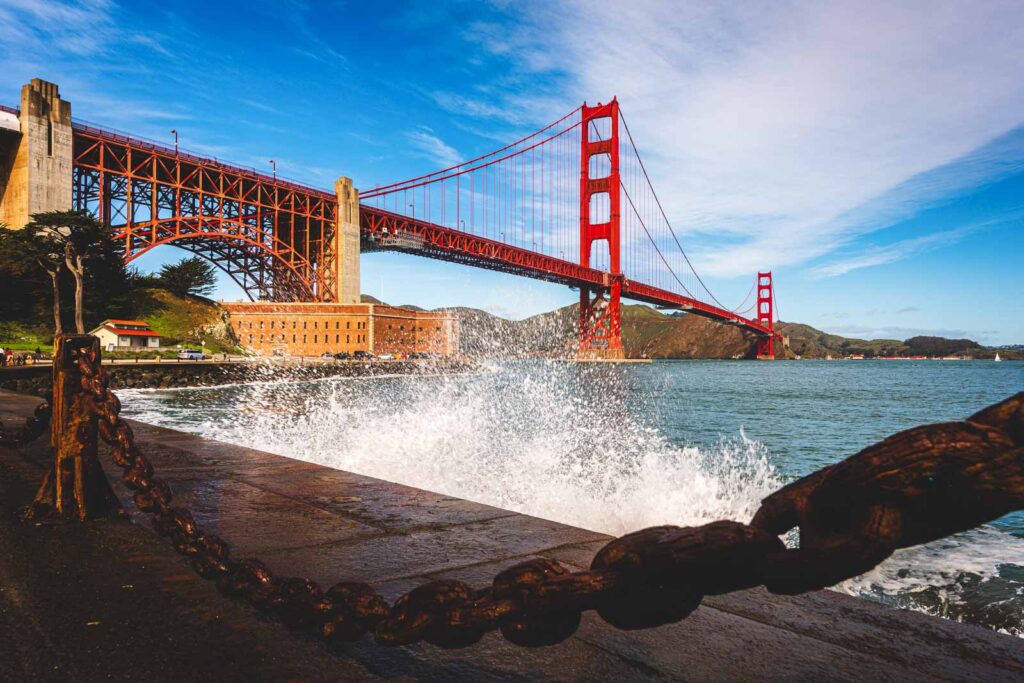 A wave from the San Francisco Bay splashing up the side of a wall at Fort Point with a view of the Golden Gate Bridge.