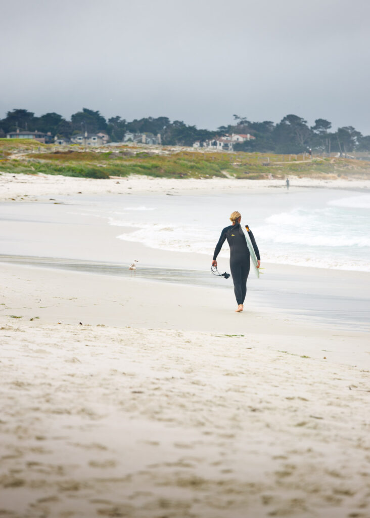 A male surfer in a wetsuit walking along Asilomar Beach on an overcast day.