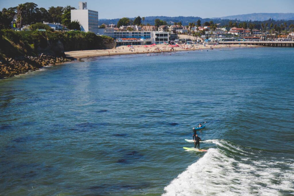 Surfers on the ocean with a kayaker besides a busy Cowel Beach in Santa Cruz.