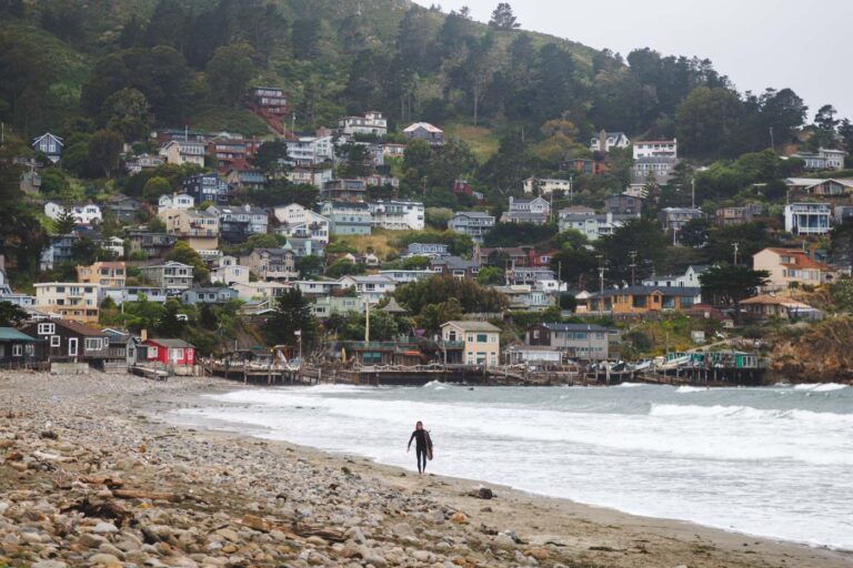 A surfer walking along Pacifica State Beach with a backdrop of houses.
