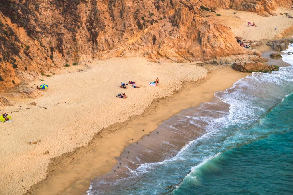 Tourists sunbathing on Gray Whale Cove State Beach next to blue ocean water and red cliffs.