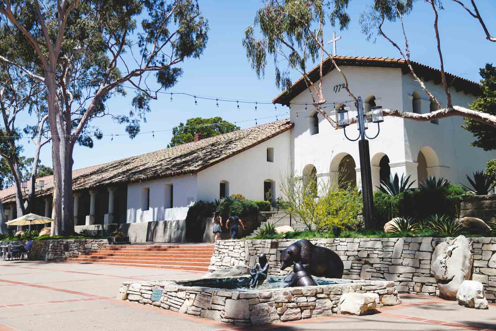 Two people entering the simple and white building of Mission San Luis Obispo de Tolosa framed by trees, walls and statues.