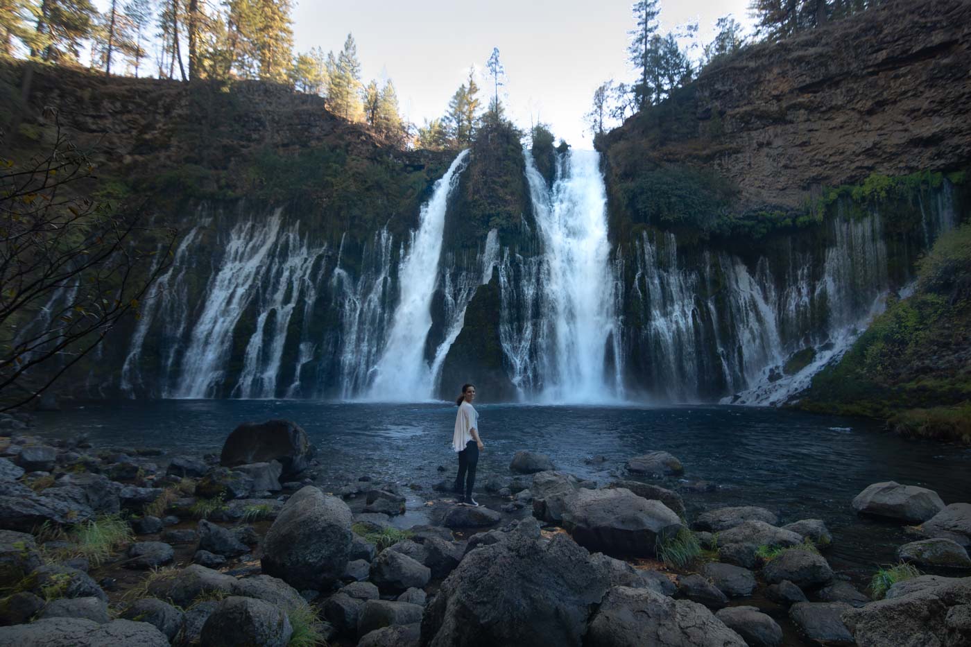 Me standing in front of Burney Falls on the rocky shores before the big blue pool of water.