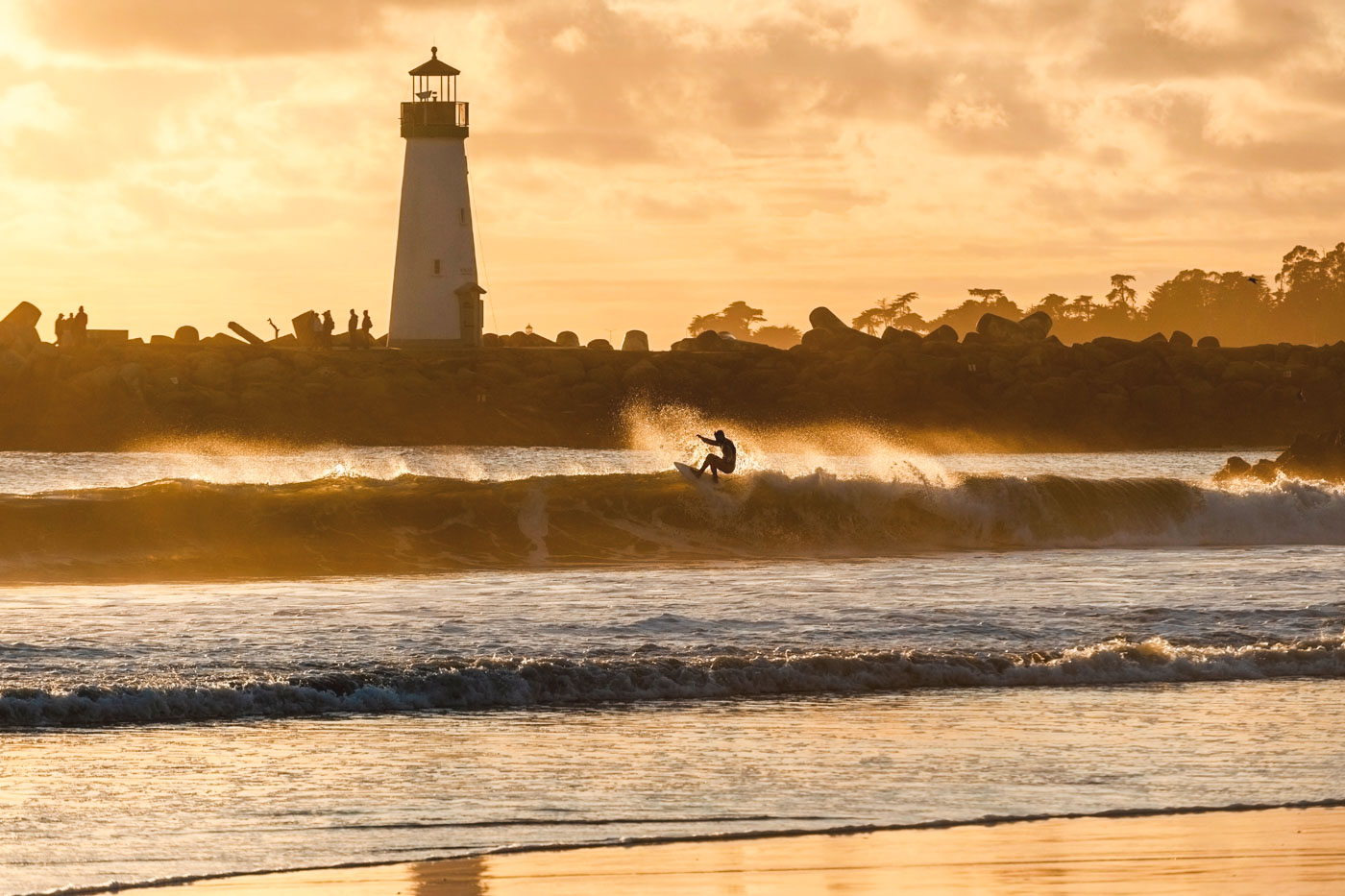 A surfer carving a wave in a golden hour glow with a backdrop of a lighthouse at Seabright Beach.