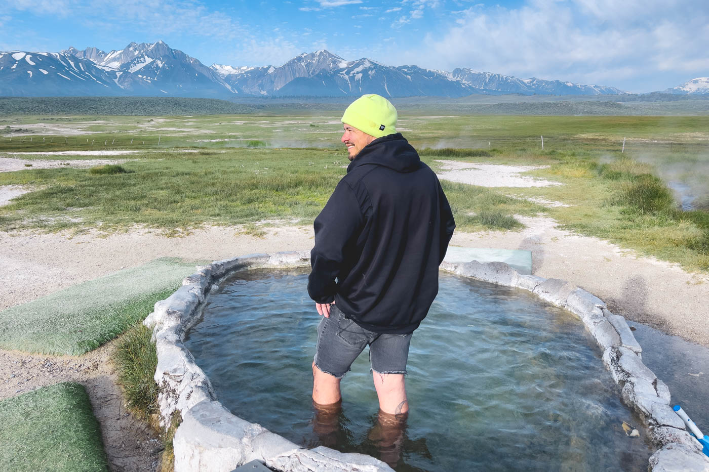 Garrett in shorts, a jumper and a bean standing in Hilltop Hot Spring with a mountain view on a sunny day.