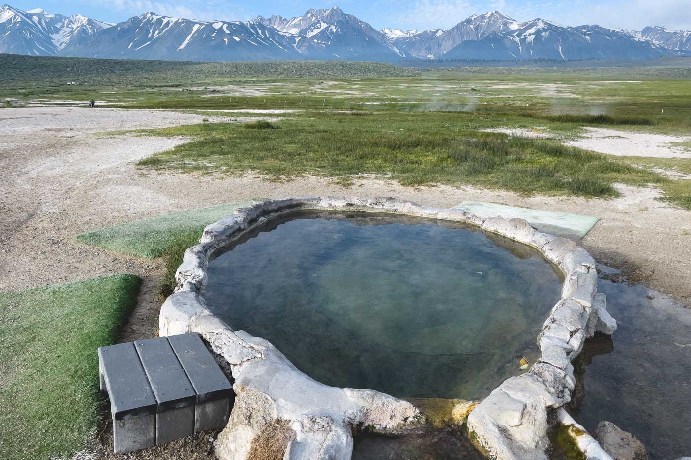 A step into Hilltop Hot Spring with a view over a field and the Sierra Nevada mountain range.