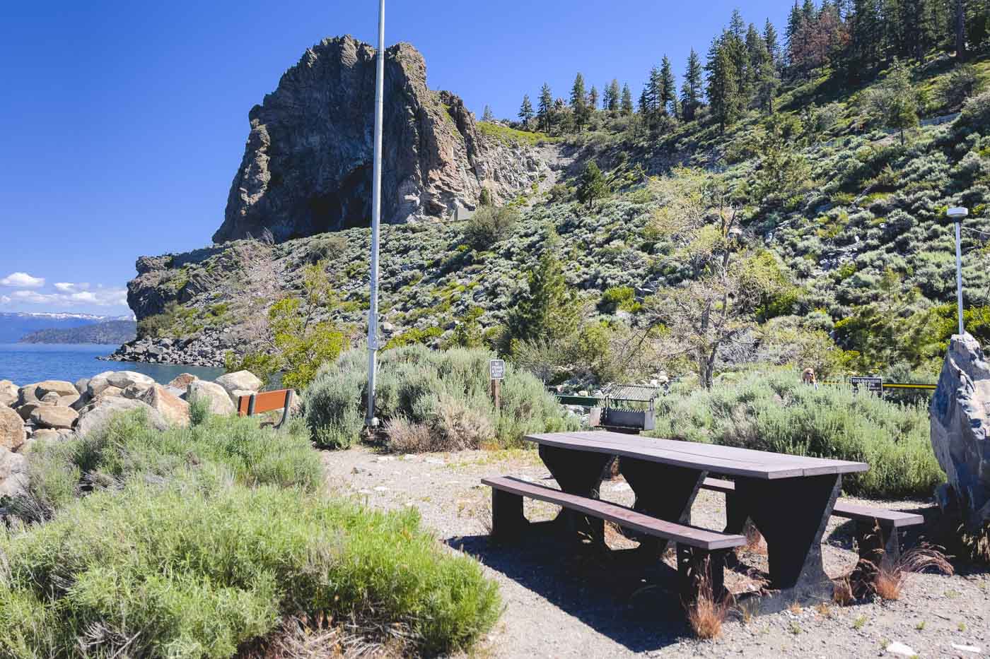 An empty picnic bench in the Cave Rock day use area with a view of Cave Rock beside Lake Tahoe in the distance.