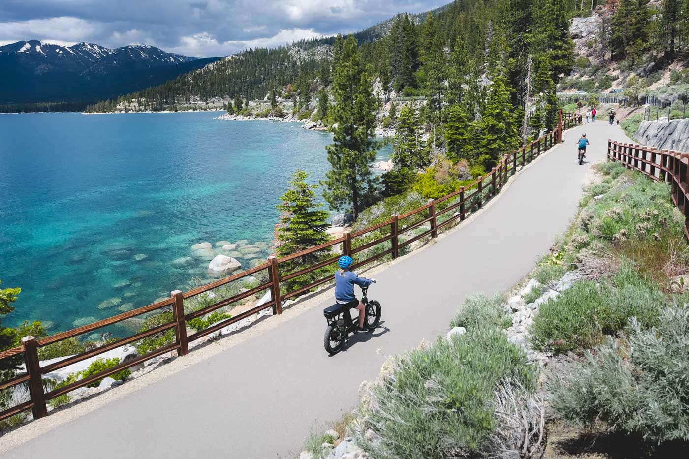 Two girls cycling along the paved pathway of the Tahoe East Shore Trail with a view of the blue water of the lake and trees along the trail.