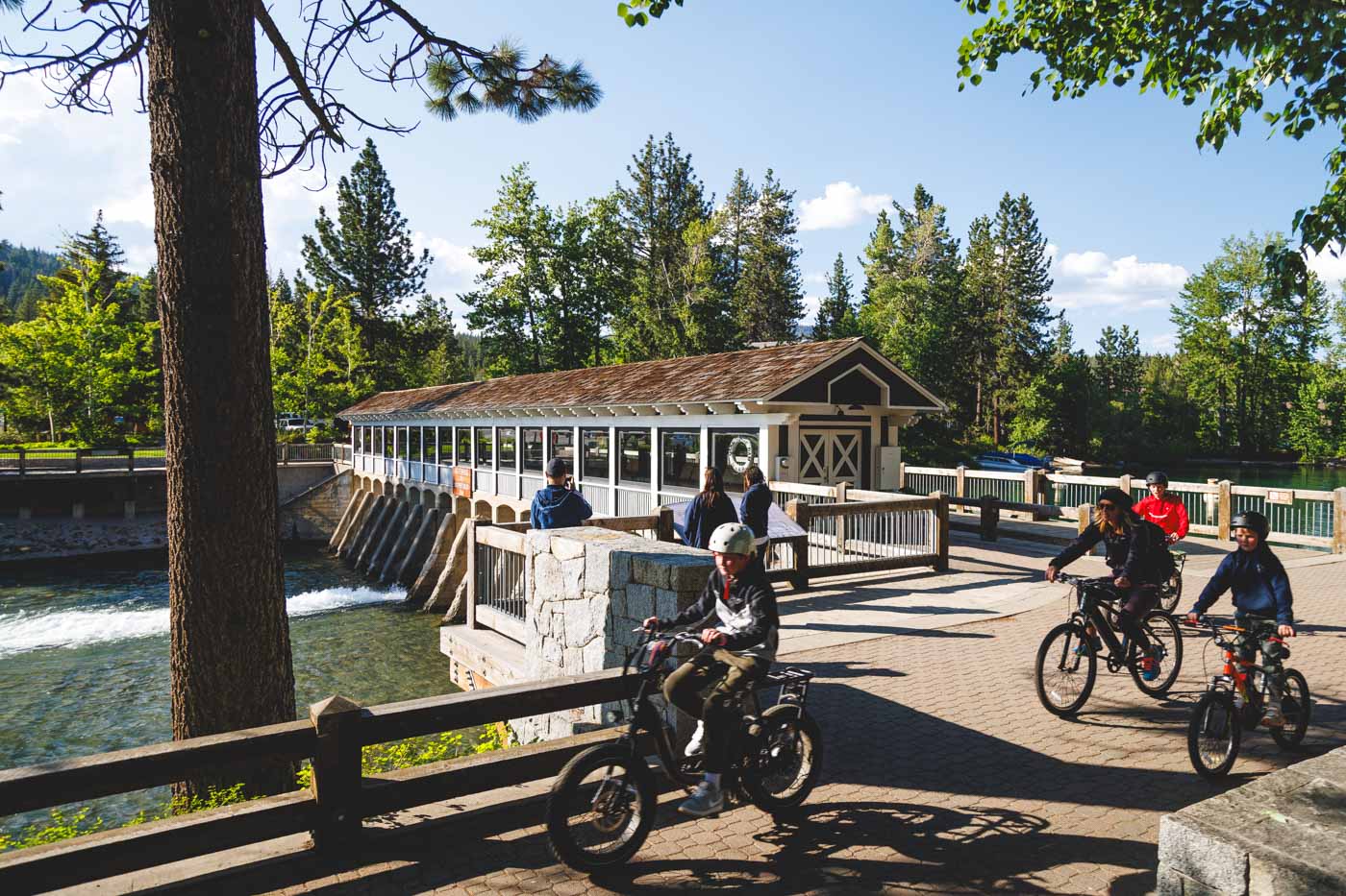 A family cycling past the bridge that sits over Tahoe Dam with trees all around.