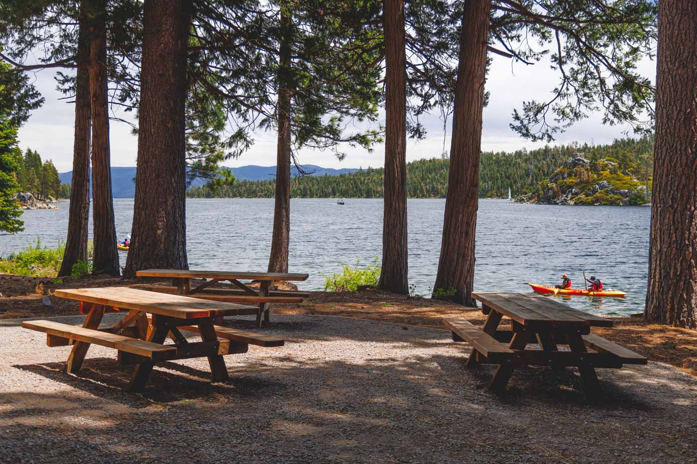 Three empty picnic tables in the day use area of Emerald Bay while kayakers row past in the background on the lake.