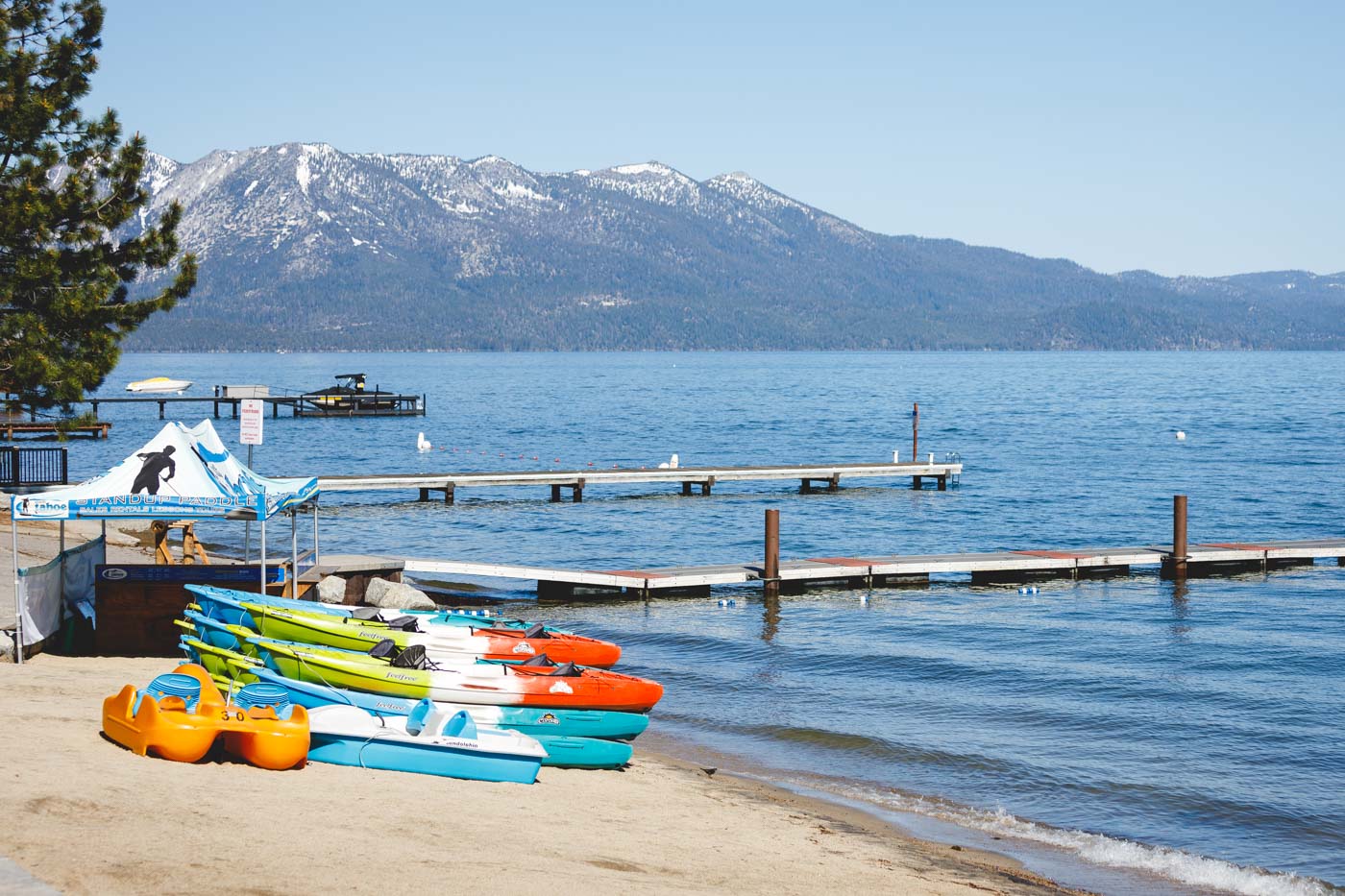 Kayaks lined up on El Dorado Beach with two jetties on Lake Tahoe and a mountain view in the distance.