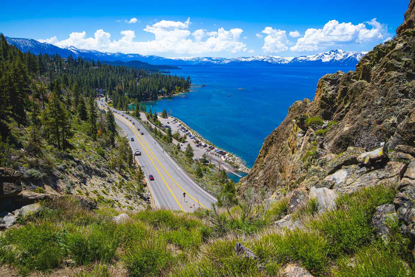 A view from Cave Rock overlooking the highway besides Lake Tahoe and snowy mountains in the distance on a sunny day.