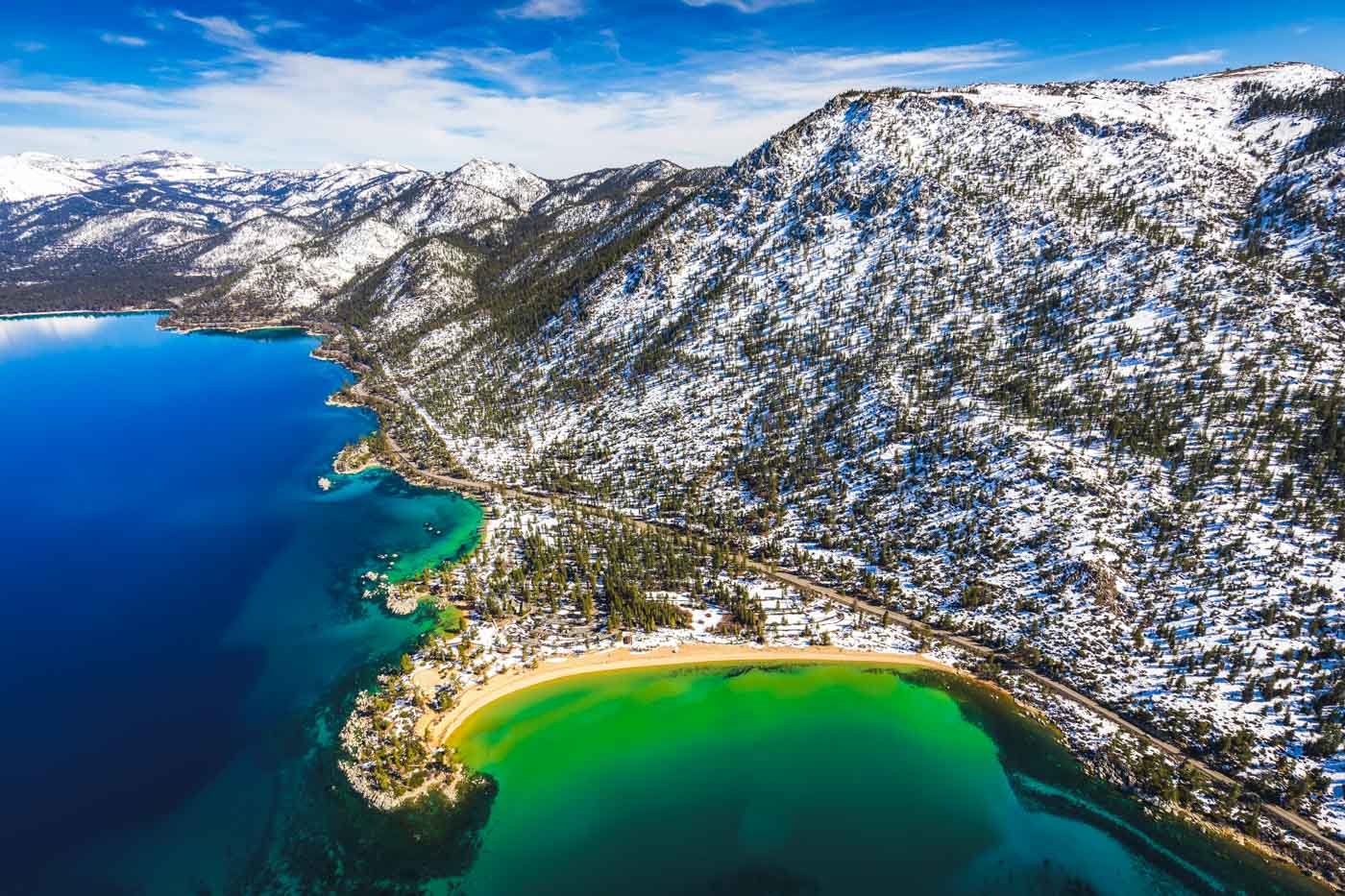 A beach along the shores of Lake Tahoe with some green water and with the surrounding area covered with snow as seen from a helicopter.