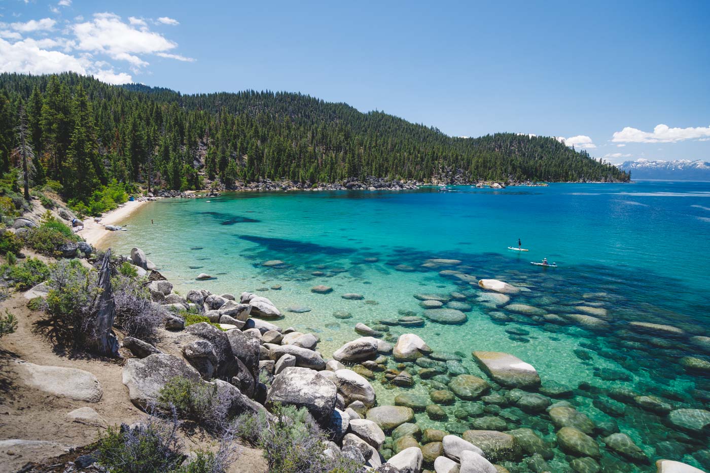 Two paddle boarders on the turquoise waters of Lake Tahoe heading towards Boaters Beach over rocks in the lake with trees around the beach.