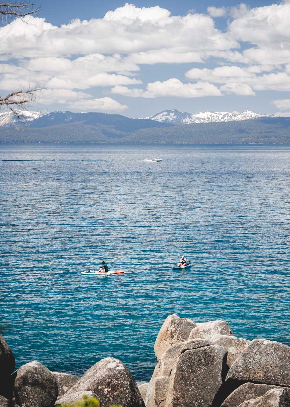 Two people paddle boarding on Lake Tahoe with a speedboat in the background and mountains and clouds in the distance.