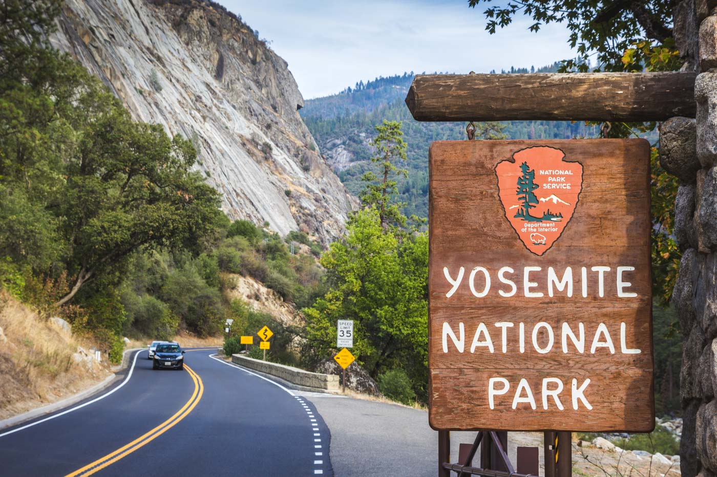 One of the many entrance signs to Yosemite National Park beside a road running past a mountain.