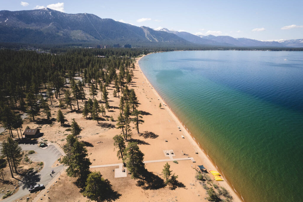 Aerial view over a huge stretch of Nevada Beach along the shore of Lake Tahoe with trees dotted around and day use areas between.