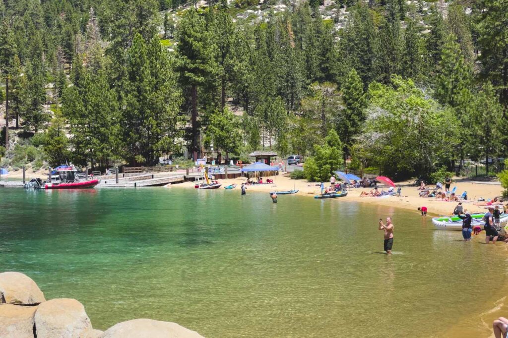 Lots of beach goers in and around Sand Harbor Beach with trees behind and a pier to the left.