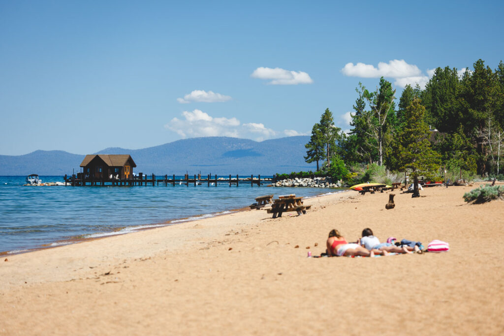 Two girls sunbathing on Nevada Beach near a copse of trees and a house on a pier on Lake Tahoe.