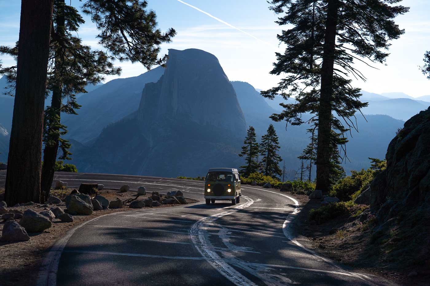 A VW Bus riding down turn in the road on Glacier Point Road with Half Dome in the background.