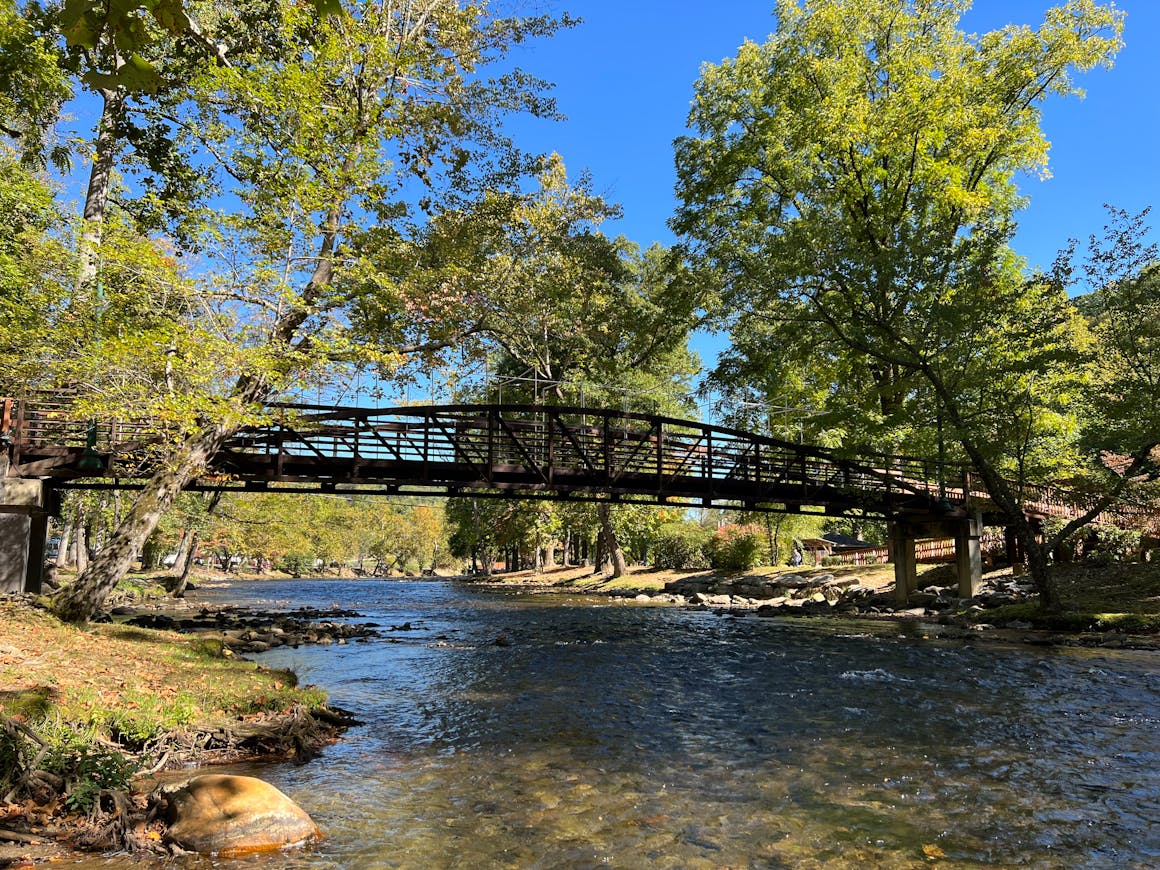 Peaceful view of a wooden bridge crossing a river in Oconaluftee Island Park, surrounded by lush trees.